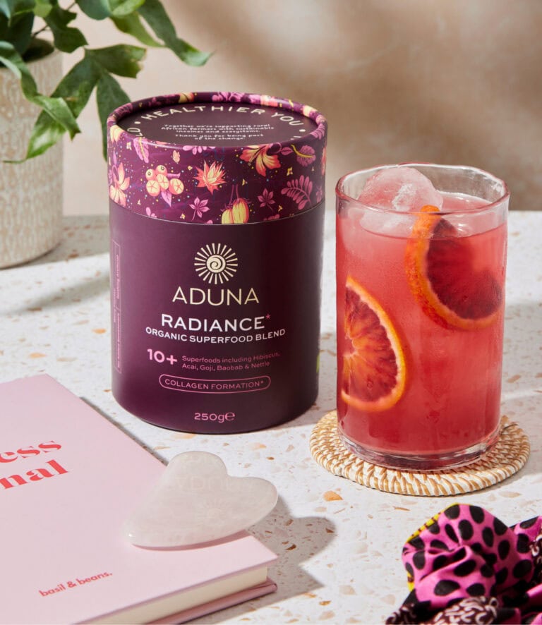 Aduna Radiance superfood blend tube in a floral purple design, beside a pink iced drink garnished with blood orange